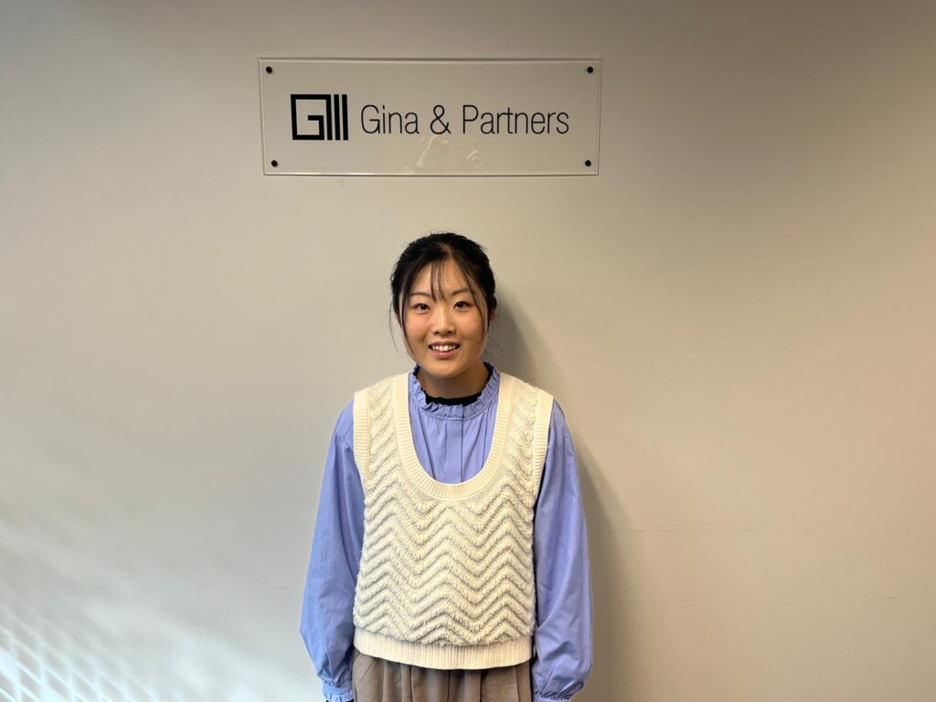 Rei さんがGina & Partnersのロゴの前で微笑む様子